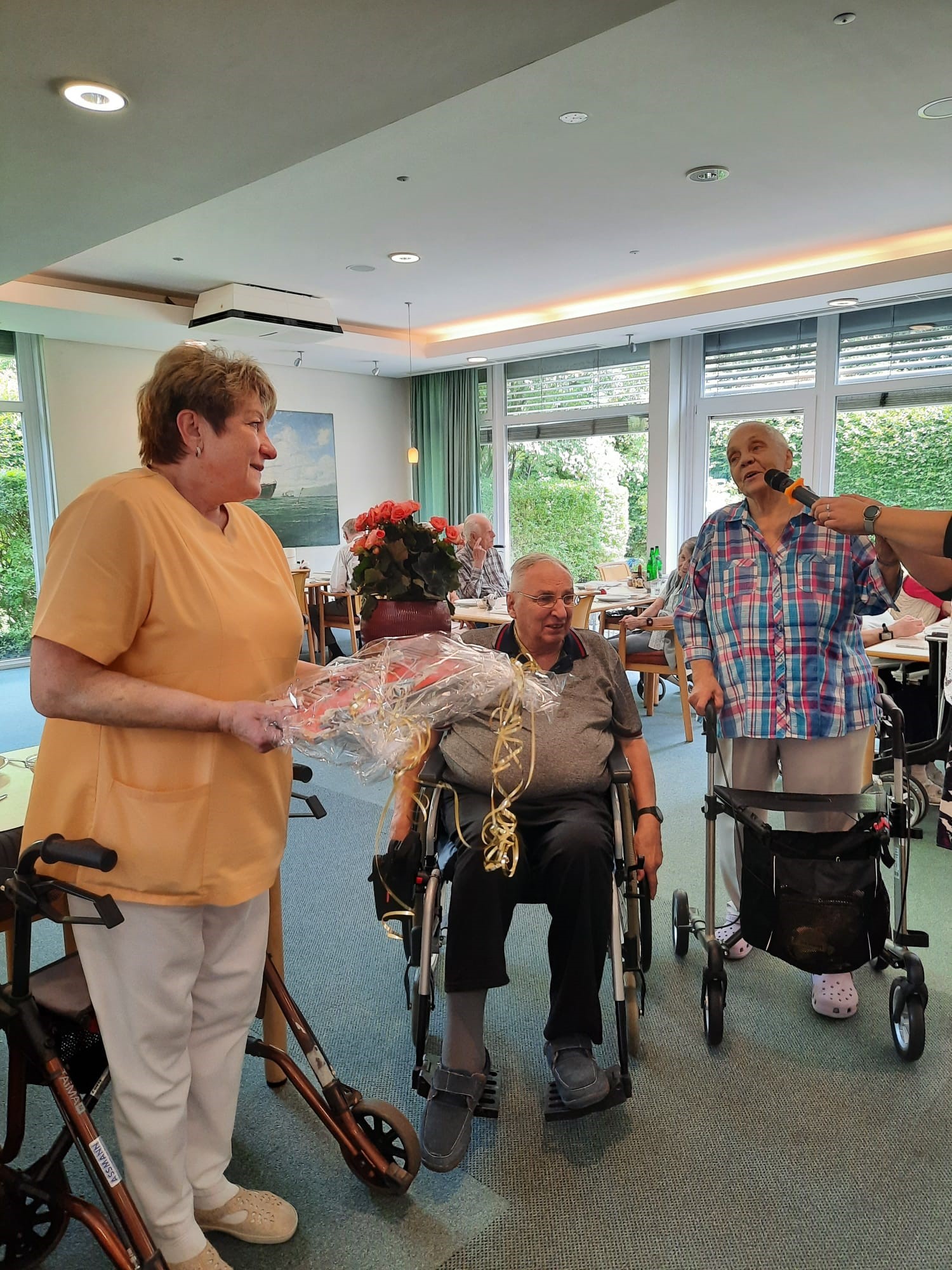 Eine Pflegefachkraft im orangefarbenen Kasack steht mit einem Geschenk in der Hand neben zwei Bewohnerinnen im Aufenthaltsraum. Einer der Bewohner sitzt im Rollstuhl, die andere Bewohnerin spricht in ein Mikrofon und steht mit Rollator. Im Hintergrund sitzen weitere Bewohnerinnen an gedeckten Tischen.